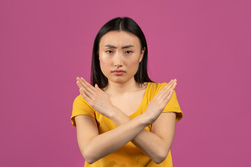 Portrait of serious asian lady showing stop gesture with crossed hands, posing over violet studio background