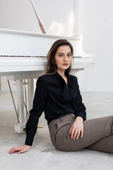 Portrait of a young beautiful female musician in a black shirt sitting on a floor with her back against the white grand piano