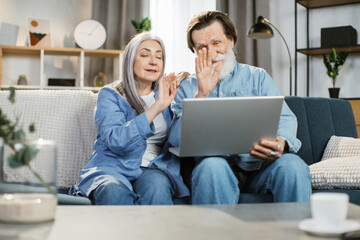 Happy mature family in casual outfit sitting together on couch and using wireless laptop. Smiling senior wife and husband spending time for communicating online with children relatives waving hands