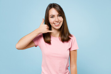 Fototapeta premium Young smiling happy caucasian woman 20s in pink t-shirt look camera doing phone gesture like says call me back isolated on pastel plain light blue background studio portrait. People lifestyle concept.