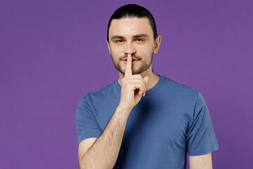 Young secret man 20s wearing blue t-shirt with blank print design say hush be quiet with finger on...