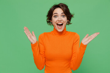 Young surprised impressed amazed shocked happy woman 20s in casual orange turtleneck spread arms hands isolated on plain pastel light green color background studio portrait. People lifestyle concept