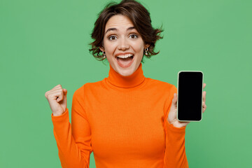 Young happy woman 20s wear casual orange turtleneck hold in hand use mobile cell phone with blank screen workspace area do winner gesture isolated on plain pastel light green color background studio.