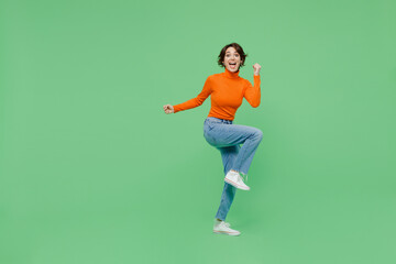 Fototapeta na wymiar Full body young woman 20s wear orange turtleneck doing winner gesture celebrate clenching fists say yes raise up leg isolated on plain pastel light green color background. People lifestyle concept