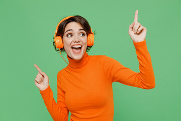 Young smiling happy cool woman 20s wear casual orange turtleneck headohones listen to music dance have fun point index finger up isolated on plain pastel light green color background studio portrait