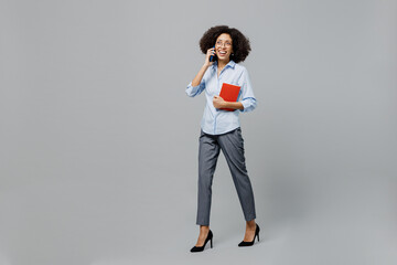 Full body fun young employee business corporate lawyer woman of African American ethnicity in classic formal shirt work in office talk speak on mobile cell phone hold book isolated on grey background.