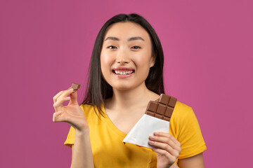 Happy korean lady eating chocolate, enjoying yummy dessert and smiling at camera, standing over violet studio background