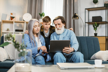 Family leisure, technology concept. Young grandson hugging his grandmother and grandfather in casual domestic clothes, sitting together on soft couch at home and watching interesting movie on laptop