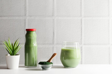 green smoothie in glass and spirulina algae powder in bowl with spoon,flower on white ceramic tile background.self care and healthy lifestyle concept.