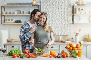 Smiling young european woman prepares salad, man with stubble hugs wife at table with organic vegetables
