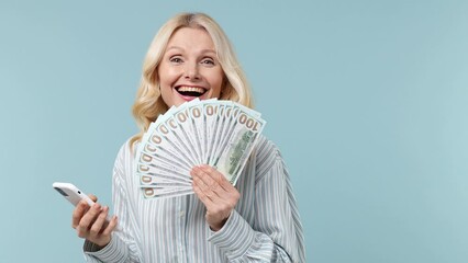 Excited joyful elderly blonde woman lady 40s years old wears white shirt use mobile cell phone hold fan of cash money in dollar banknotes isolated on plain pastel light blue background studio portrait