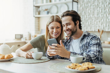 Smiling young european family look at smartphone, chatting in social networks, drink tea in modern kitchen