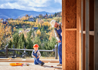 Father with toddler son building wooden frame house. Male builders hammering nail into plank on...