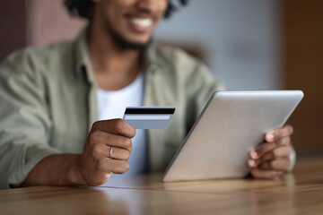 Cropped happy millennial african american guy with tablet sitting at table and hold credit card to pay