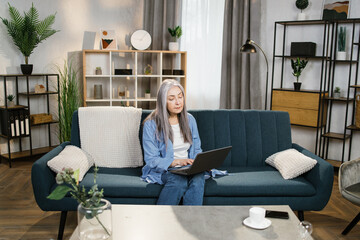 Online education and distance work from home. Happy smiling senior woman, sitting on blue sofa and studying with laptop at home, while watching webinar or online lessons.