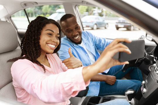 Happy black couple driving car and taking selfie on smartphone