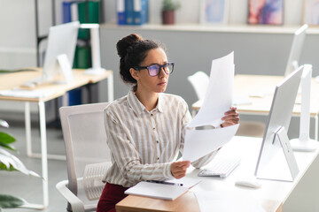 Concentrated latin businesswoman working with documents and reports, sitting at workplace in office interior