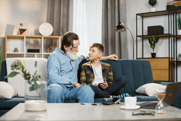 Happy grandfather with grey beard cuddling smiling small child boy playing online mobile game...