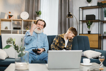 Positive elder man and his charming grandson sitting together on couch and using wireless joysticks for playing video games. Lovely different age family spending free time with fun at home.