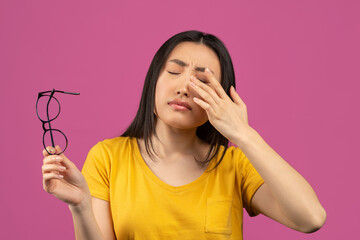 Young asian lady suffering from eye pain, taking off glasses and touching her eye, standing over...