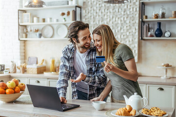 Happy young european husband with stubble and wife shopping online and show credit card in modern...