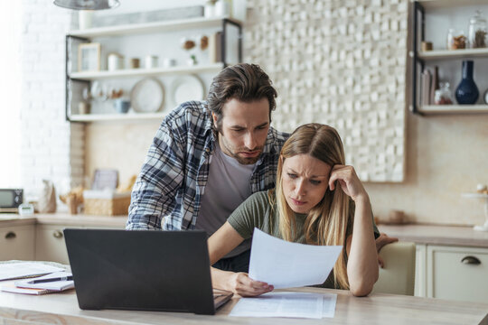 Unhappy Young Caucasian Family Think, Look At Document, Pay Bills And Taxes At Table With Laptop