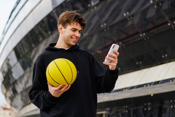 Young smiling basketball fan in black hoodie holding ball and watching match on smartphone. Happy man win a bet.
