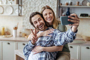 Satisfied young european lady hugging male with stubble, guy shows peace sign, makes selfie on phone