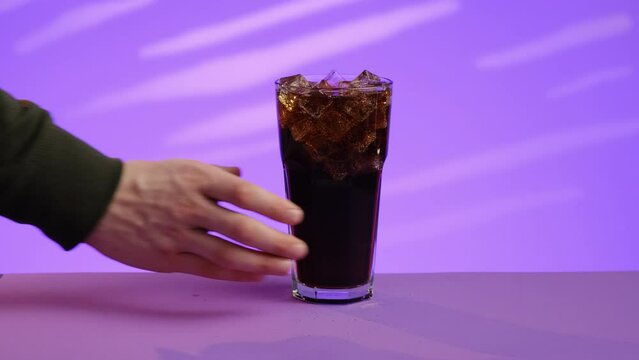 A glass with a drink and ice on a purple background. A hand in green clothes takes a glass and takes it over the edge of the frame
