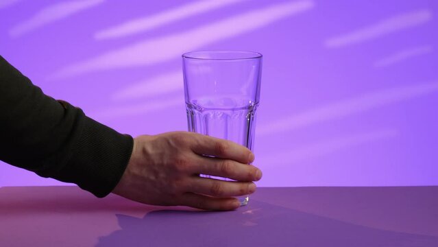Transparent glass glass on a purple background. A hand in green clothes takes an empty glass and takes it over the edge of the frame