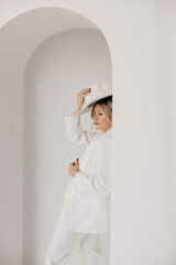 Confident young blond woman smiling, looking at camera isolated on white background. Studio portrait of successful friendly female in white suit and hat, posing over white wall.