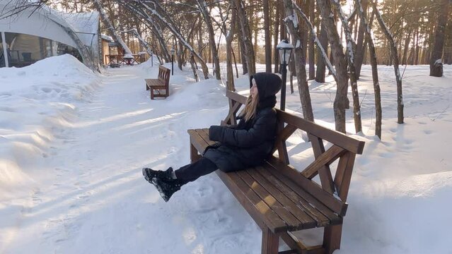 Smiling teenager girl with blone flowing hair in black coat and white hat sit on brown wooden bench in winter snow covered park