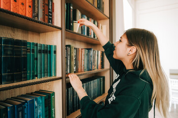 Young caucasian woman standing near bookshelf in library and choosing book for reading.