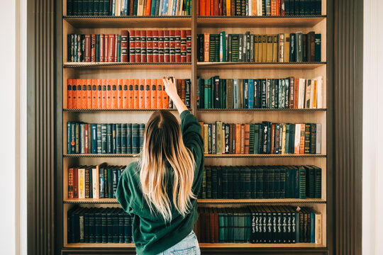 Young caucasian woman standing near bookshelf in library and choosing book for reading.