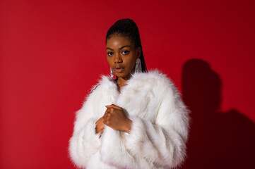 Fashionable confident Black woman wearing luxury white faux fur coat, trendy long earrings with rhinestones,  posing on red background. Copy, empty space for text