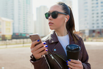 Fototapeta na wymiar Close up portrait of stylish european girl with dark hair wearing sunglasses and jacket is holding smartphone and looking aside in sunlight on city background