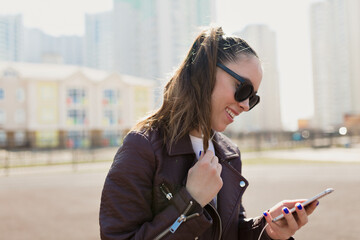 Fototapeta premium Close up portrait of incredible lovely girl with dark hair is looking at smartphone and listening music on city background in sunlight