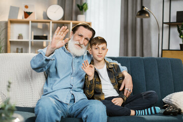 Happy grandfather sit on couch enjoy spending time with school age grandson at home, loving smiling grandparent and little boy child relax on sofa have close tender moment looking at the camera