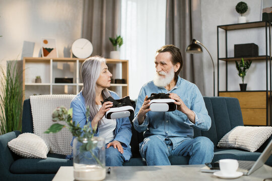 Beautiful mature woman and her bearded husband sitting together on couch with VR headset at home. Happy senior family spending leisure time at home and playing virtual reality games.