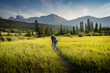 Two children running down a trail across a grassy meadow holding fishing rods in the Canadian Rockies near Crowsnest Pass Alberta Canada during summer.
