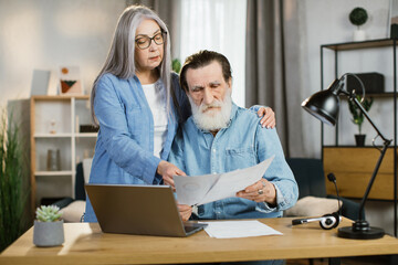 Beautiful mature woman and bearded man in casual outfit using laptop during working together. Pleasant old wife and husband business partners cooperating at bright office or working online from home