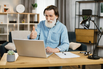 Fototapeta na wymiar Handsome caucasian senior software developer in casual wear typing on laptop while sitting at workplace and showing thumb up. Busy office worker mature gentleman using modern gadgets for work
