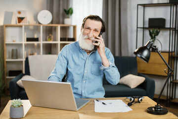 Pleasant mature caucasian businessman talking on mobile and working on laptop at table. Senior male in casual wear sitting at workplace and using modern gadgets.