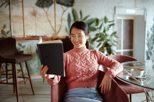 A cheerful young asian woman reading a book while drinking a coffee in a cafeteria.