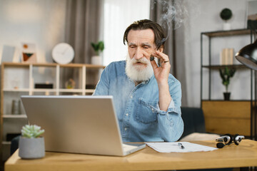Portrait of handsome elegant mature business man with cigar sitting at desk with laptop during video call and looking at camera. Senior man using modern devices for online conversation at work.