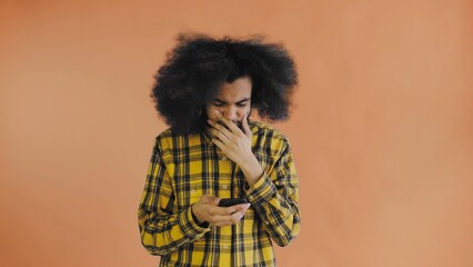 A young man with an African hairstyle on an orange background looks at the phone and is happily surprised. Emotions on a colored background