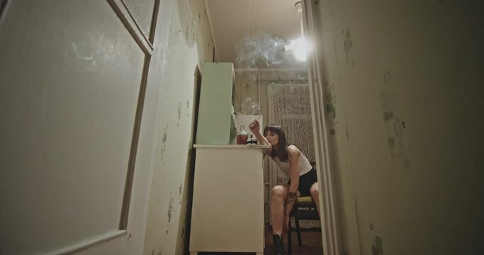 Female addict smoking weed in kitchen. Low angle zoom in view of high woman leaning on table and smoking cannabis joint in grungy kitchen at home