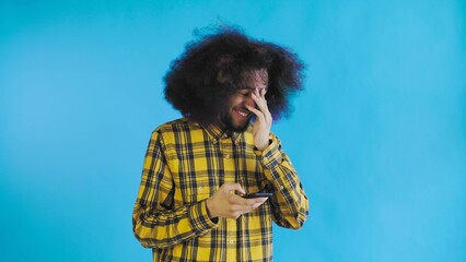 A young man with an African hairstyle on a blue background looks at the phone and happily texts with someone. Emotions on a colored background