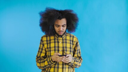 A young man with an African hairstyle on a blue background looks at the phone and doubts. Emotions on a colored background