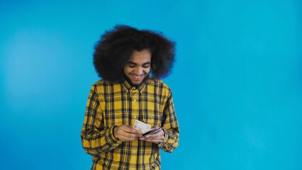 A happy African-American man holding banknotes in his hands, kissing them and looking at the camera, stands isolated on a blue background